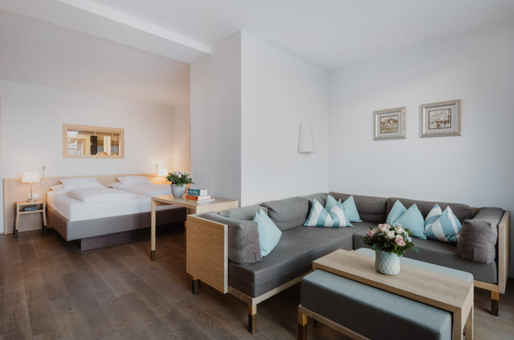 Room Moosham in the Elixhauser Wirt in Salzburg with large gray corner couch with light blue cushions, tables with flowers, white double bed and wooden floor.